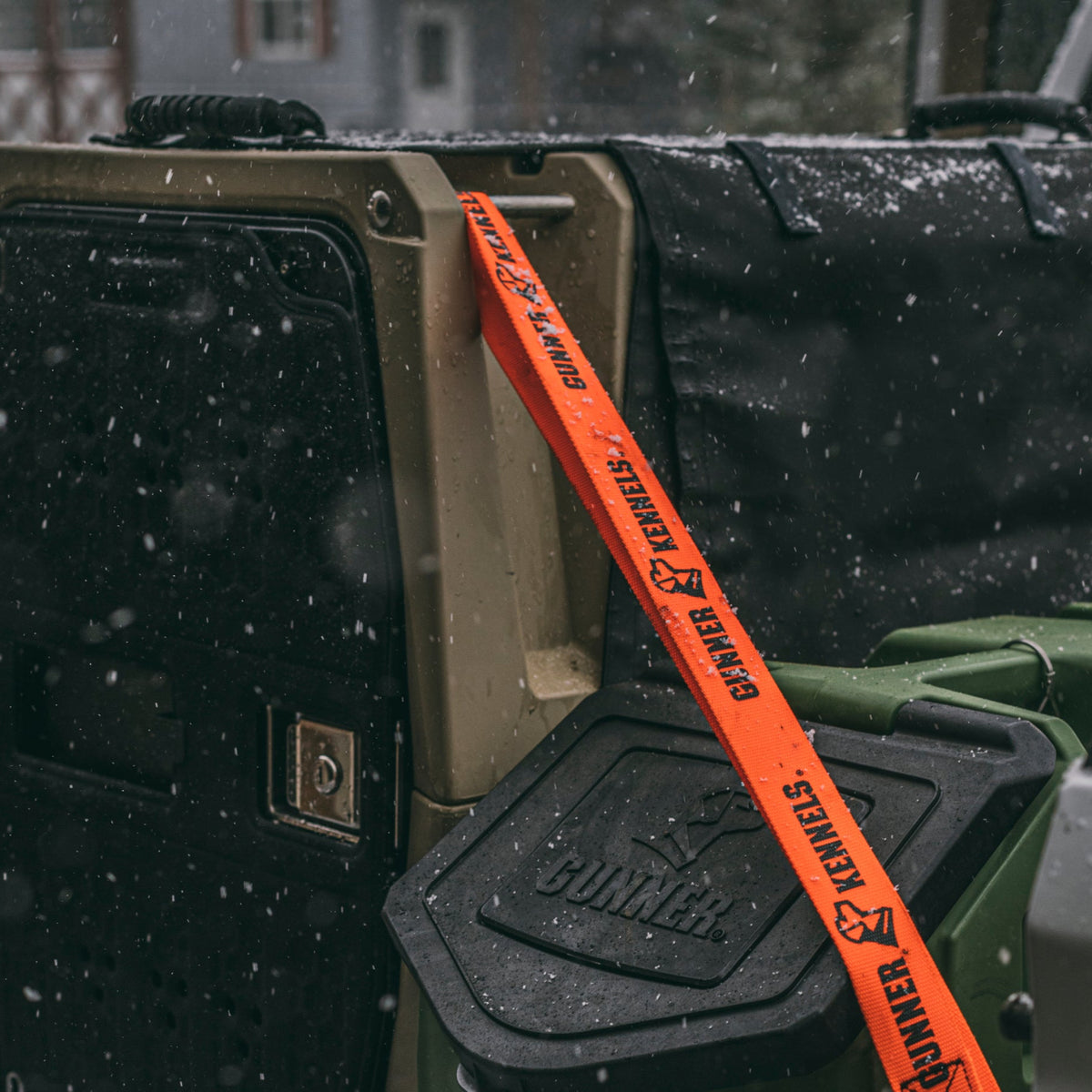 An orange strap featuring the GUNNER logo is attached to a rugged outdoor container in a snowy environment, highlighting the resilience of the ALL-WEATHER KIT 2.0 BLACK by GUNNER. Snowflakes fall gently around, with a black bag positioned nearby.