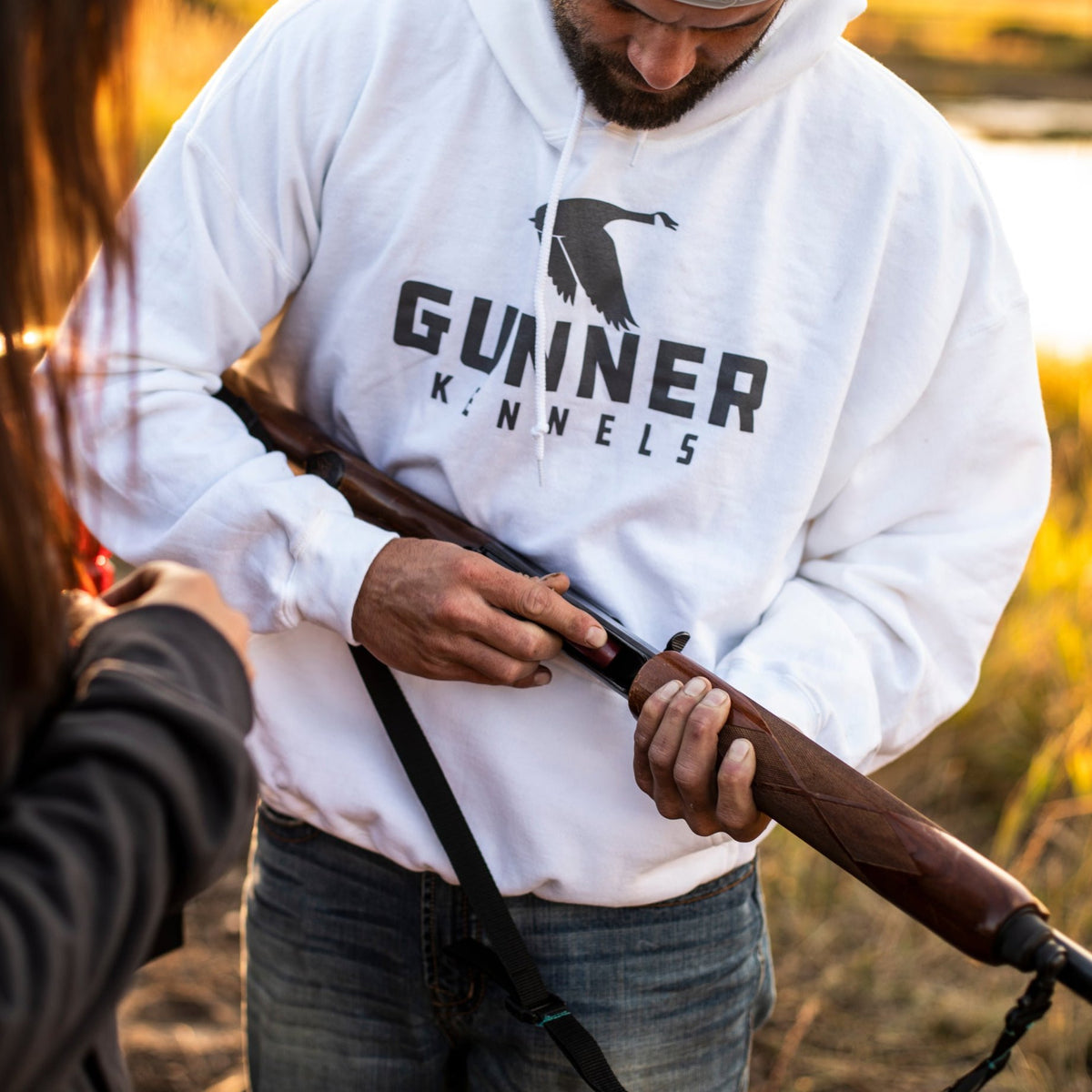 A person in a white GUNNER OG HOODIE is loading or checking a rifle outdoors with another standing nearby, surrounded by grass and sunlight.