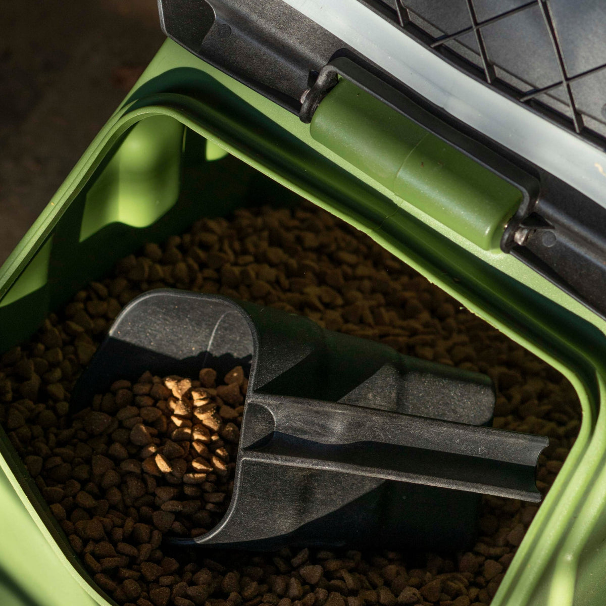A close-up of a GUNNER FOOD SCOOP inside a green container filled with dry pet food. The open lid lets sunlight reveal the pellets and the durable scoop resting inside.