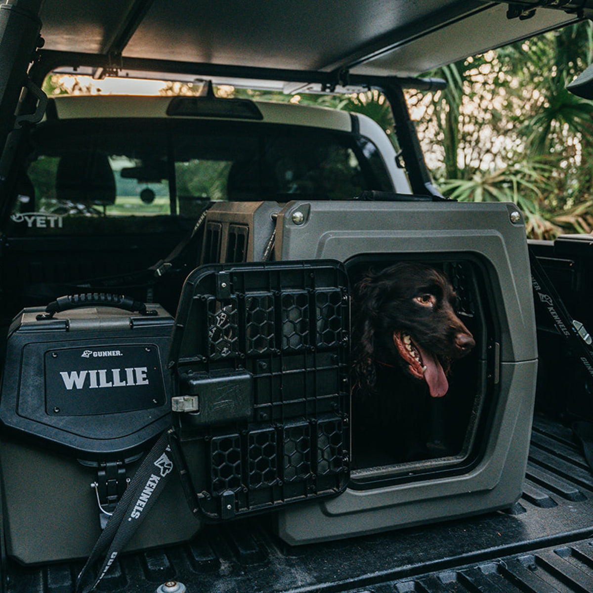 A brown dog with its tongue out sits inside a GUNNER Food Crate with a custom FOOD CRATE NAME PLATE labeled WILLIE, in the back of a pickup truck parked outdoors among green trees.