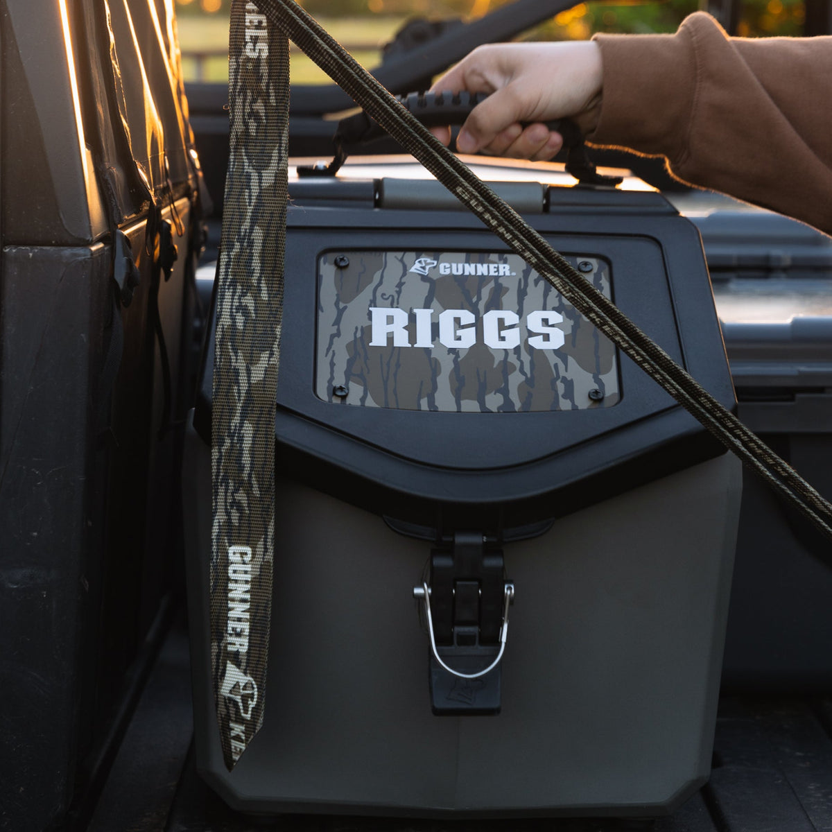 A hand grips the handle of a durable, two-tone GUNNER Food Crate with a FOOD CRATE NAME PLATE labeled “RIGGS” and a camo leash attached, all placed in the back of a vehicle.