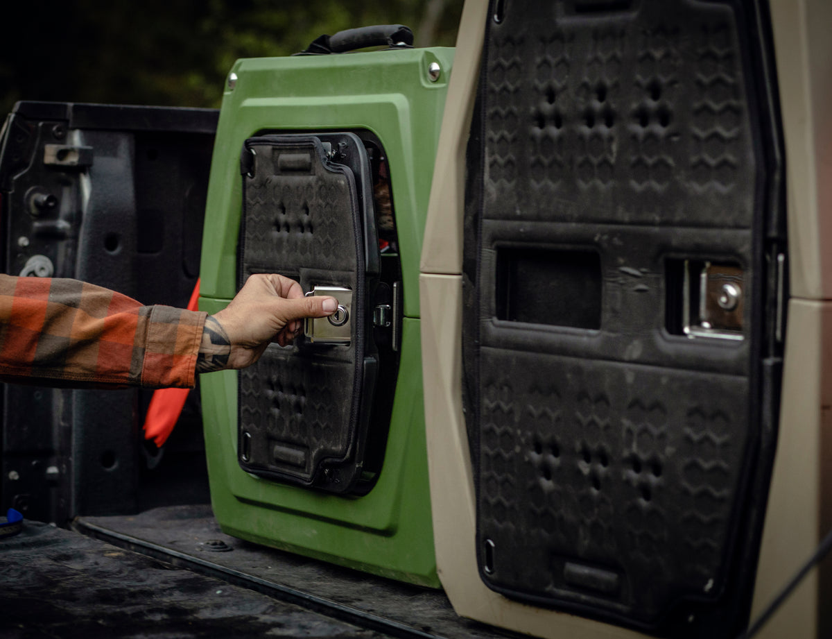 Wearing a plaid shirt, someone opens the latch of a green storage case with thermoformed foam in a truck bed, next to a larger beige GUNNER kennel and the GUNNER COLD WEATHER DOOR COVER.