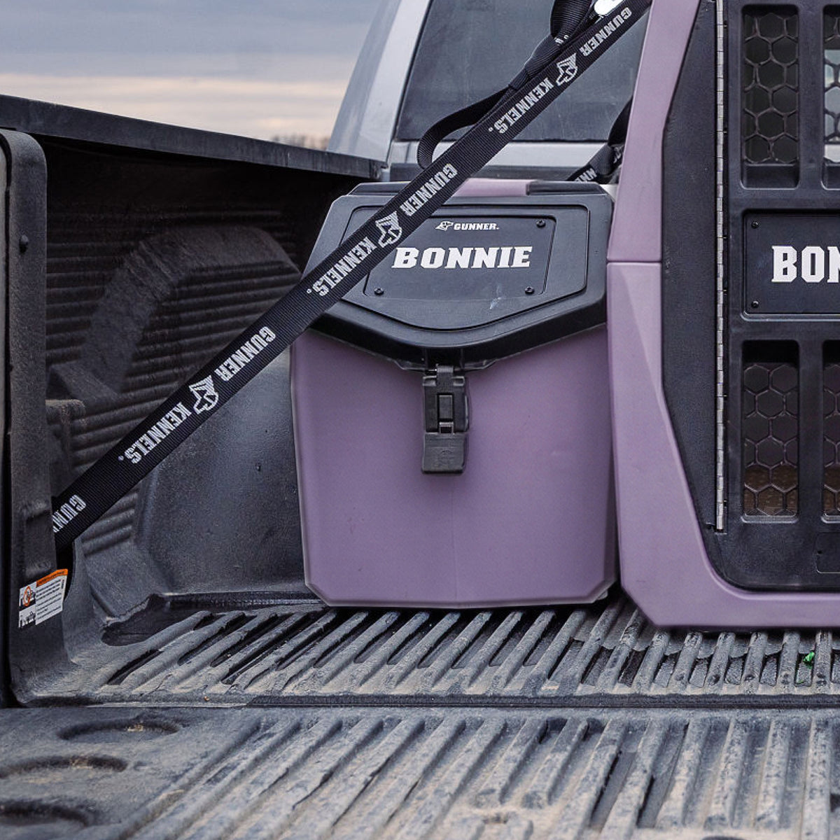 A purple BONNIE-labeled storage container sits in the back of a pickup, secured with a black GUNNER strap beside a GUNNER FOOD CRATE - DUSK, both ready for the small run before last light.