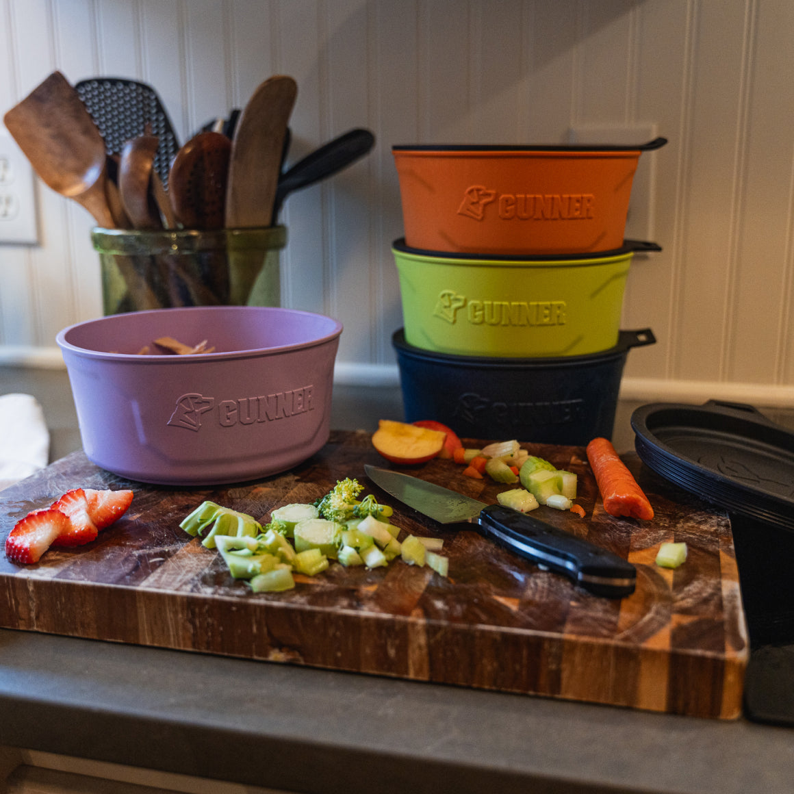 A wooden cutting board with fruits, veggies, two knives, and three stacked GUNNER DOG BOWL - LAVENDER bowls by GUNNER on a kitchen counter. Utensils sit in a glass jar; subtle lavender decor accents the background.