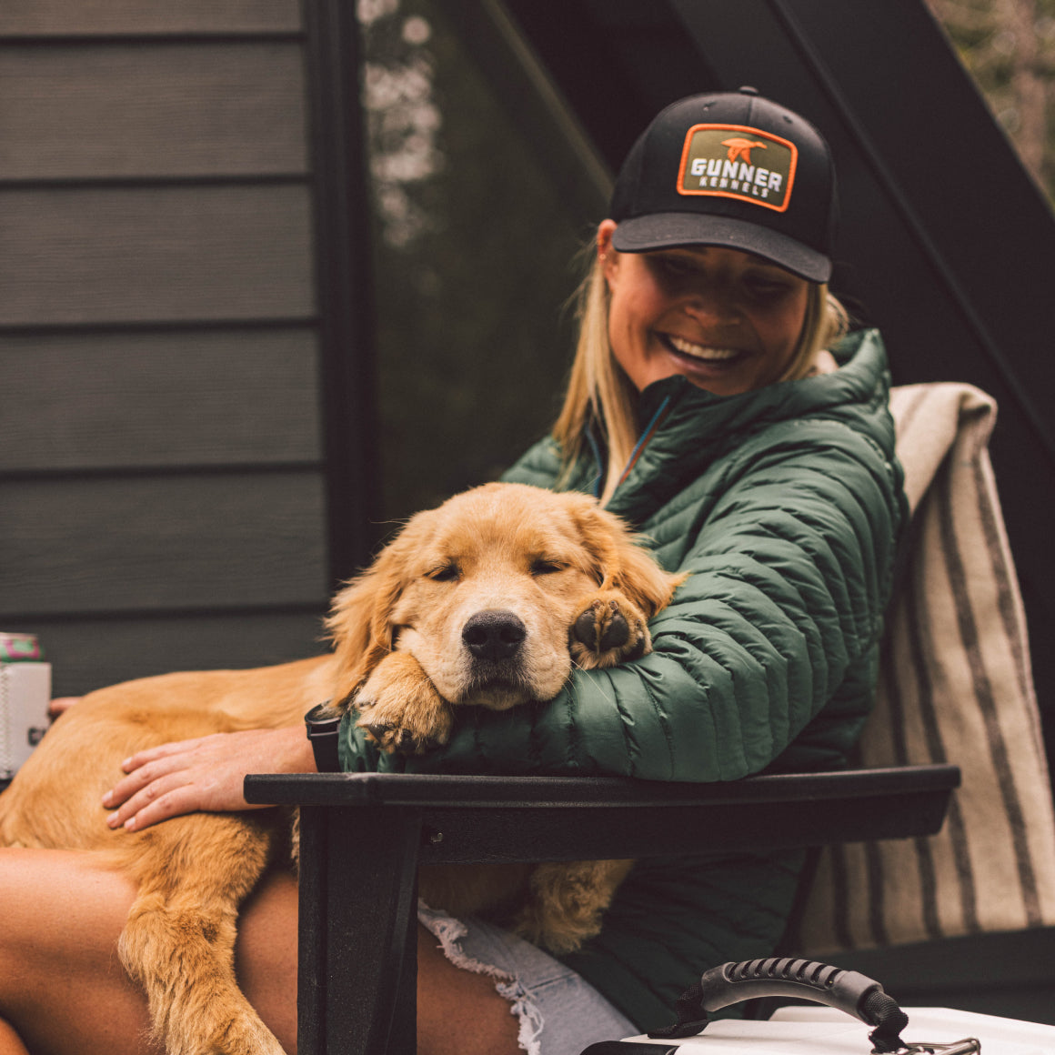 A woman in a green jacket smiles outdoors with a golden retriever puppy on her lap. She wears the GUNNER BLACK MALLARD TRUCKER hat and sits on a patio chair with a striped cushion.