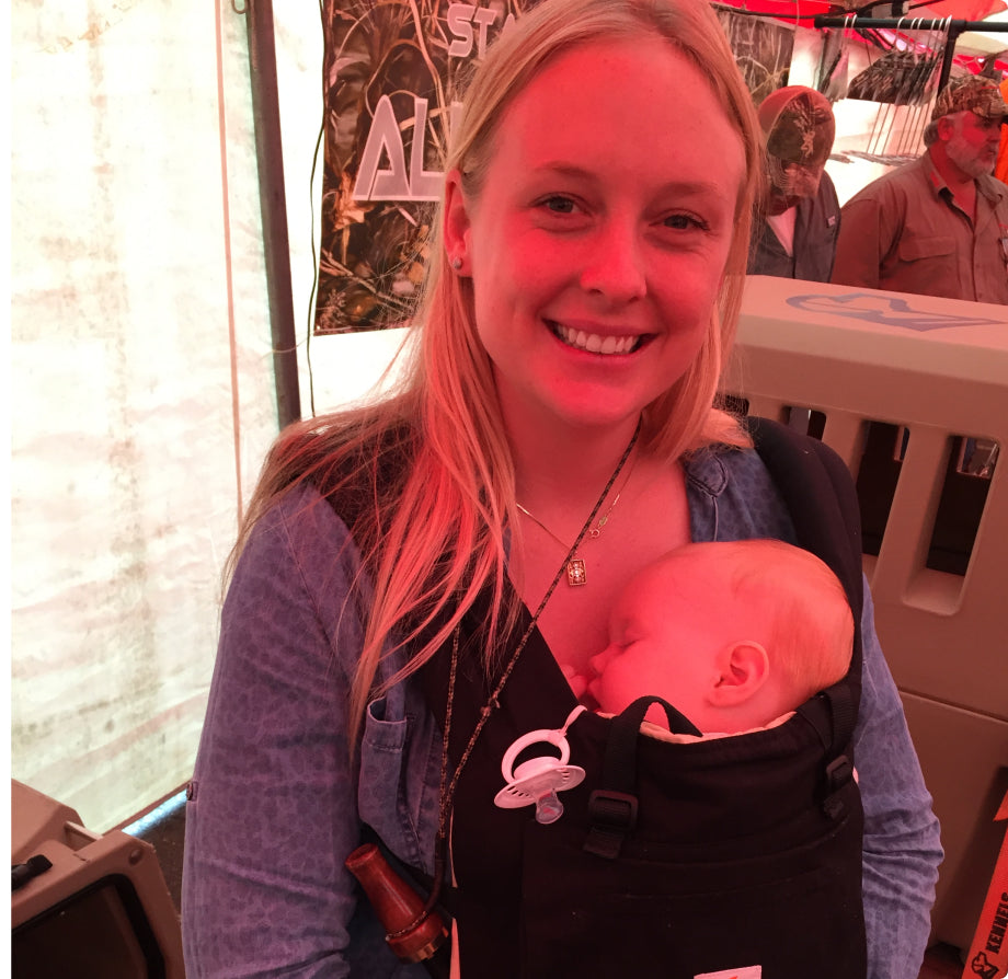 A smiling woman with long blonde hair stands indoors, wearing outdoor gear and a baby carrier on her chest that holds a sleeping infant. The woman is casually dressed, and there are people and banners in the background.