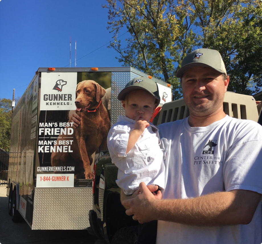 A man holding a young child stands in front of a trailer advertising Gunner Kennels, featuring a large dog image and kennel safety branding. Both wear gray hats and white shirts. Trees and blue sky frame this dog kennel scene.