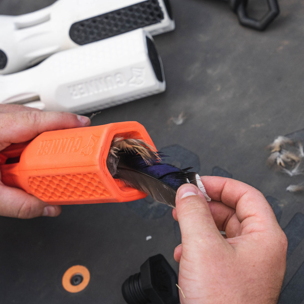 A person inserts bird feathers into an orange, textured GUNNER BUMPER by GUNNER to improve dog training, with white bumpers visible in the background on a dark surface.