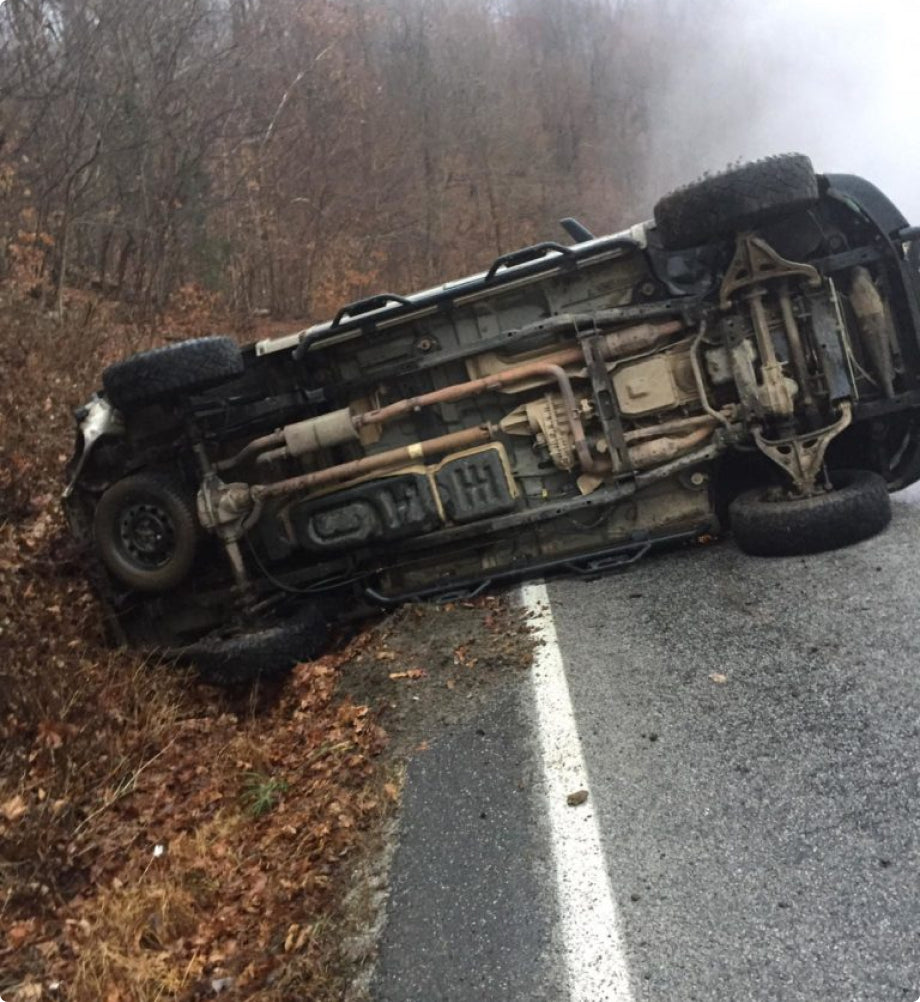 A black SUV lies flipped on its side off a wet, rural road, with smoke rising in the background. A dog crate is visible nearby among the leafless trees lining the scene.