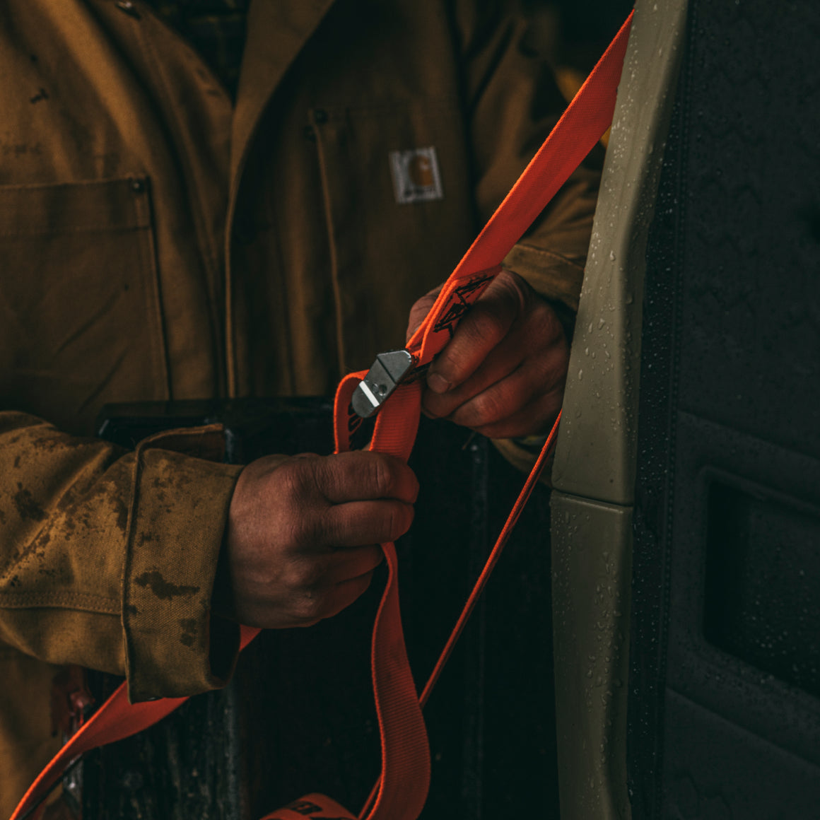Wearing a brown jacket, a person hand-tightens GUNNER TIE-DOWN STRAP KIT—crash test certified—around a vehicle as rain droplets dot the surface.