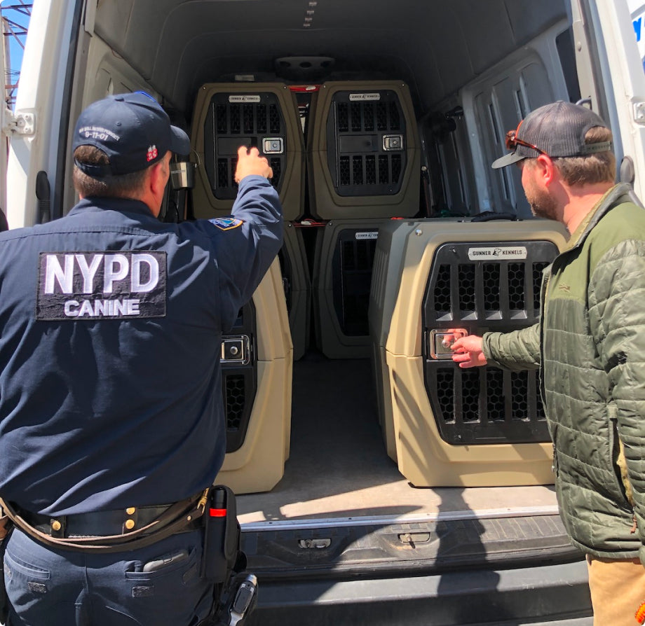 An NYPD canine officer and another man stand at the back of a van, pointing toward several large dog crates stacked inside, likely for transporting working dogs and their essential outdoor gear.