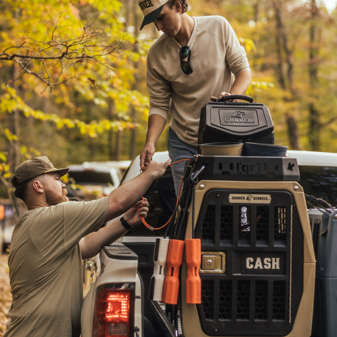 Two men stand at the back of a pickup truck in a wooded area. One hands an orange cord to the other. The truck holds a large labeled dog kennel for their hunting dog, surrounded by autumn trees in the background.
