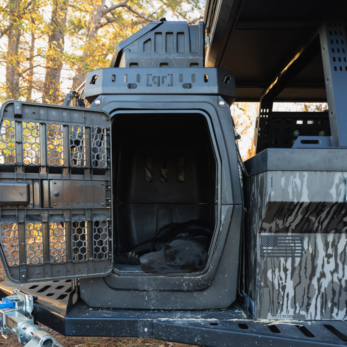 A black working dog is lying inside a ventilated dog crate attached to the back of a camouflaged vehicle, with the crate door open and trees visible in the background.