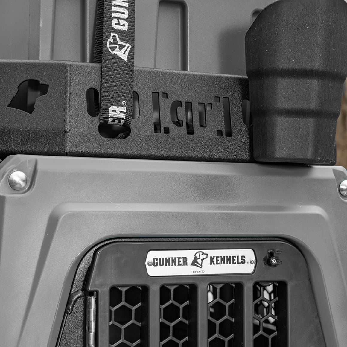 Close-up of a Gunner Kennels dog crate, showing the brand logo on a metal plate, black mesh door, and a securing strap attached to the sturdy, gray plastic and black textured exterior—perfect for any working dog kennel setup.