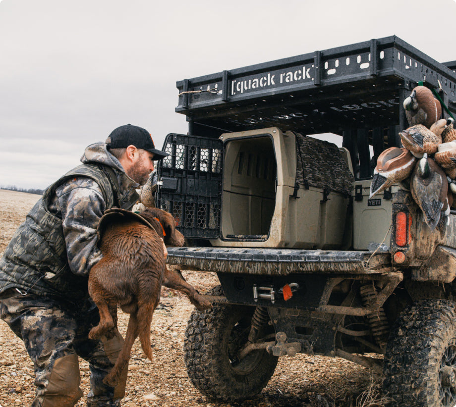 A man in camouflage lifts a wet brown working dog onto the back of an off-road vehicle loaded with hunting gear and crates. The rocky ground and overcast sky complete this rugged outdoor scene.