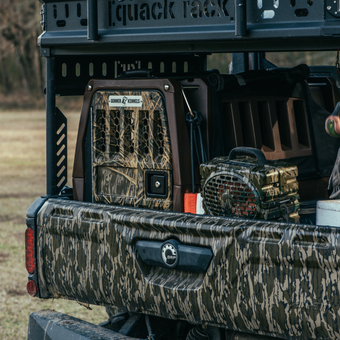 A camo truck bed holds the GUNNER G1™ KENNEL - SUSQUEHANNA/MOSSY OAK SHADOW GRASS, a camo bag, and outdoor gear—ideal for duck and goose hunters—set in front of a grassy field with trees.