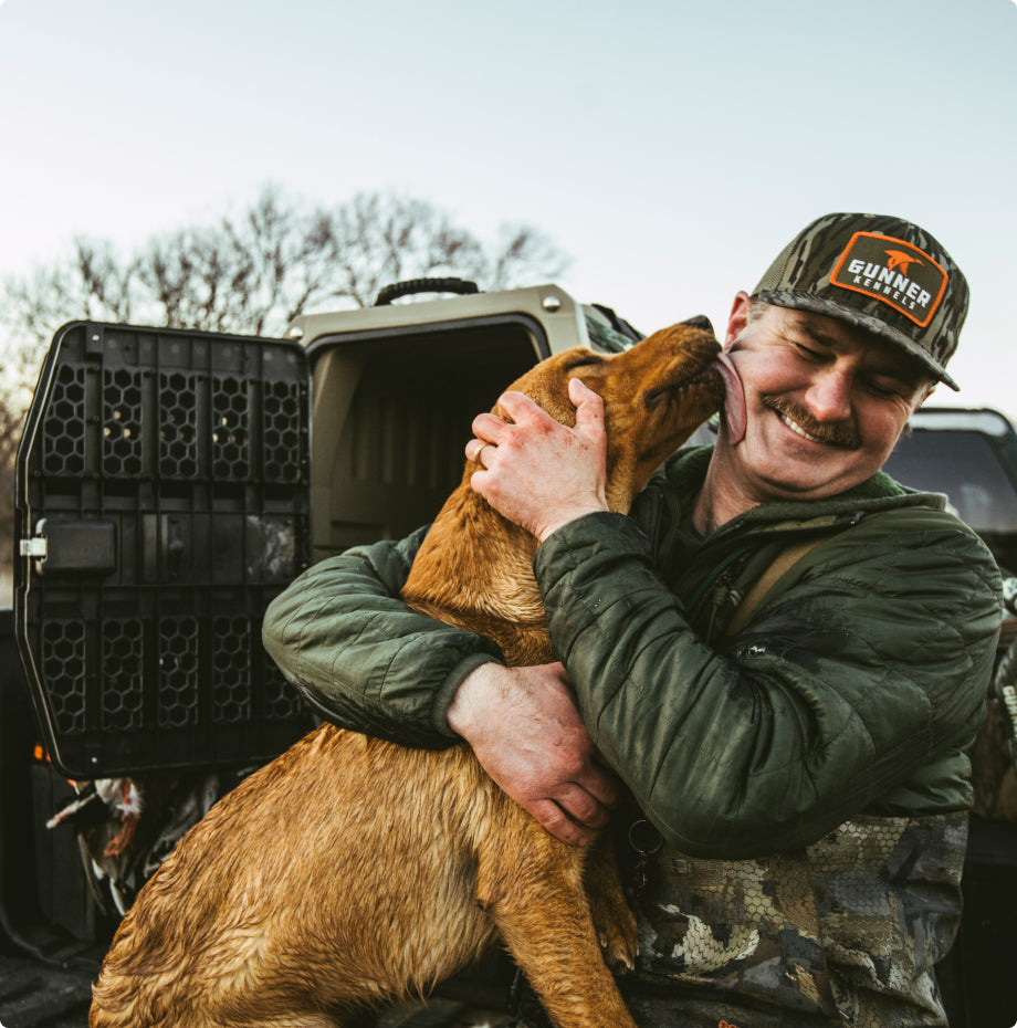 A man in outdoor gear smiles as a brown dog affectionately licks his face. He hugs the dog beside an open vehicle with a dog kennel in the background, while leafless trees stand behind them.
