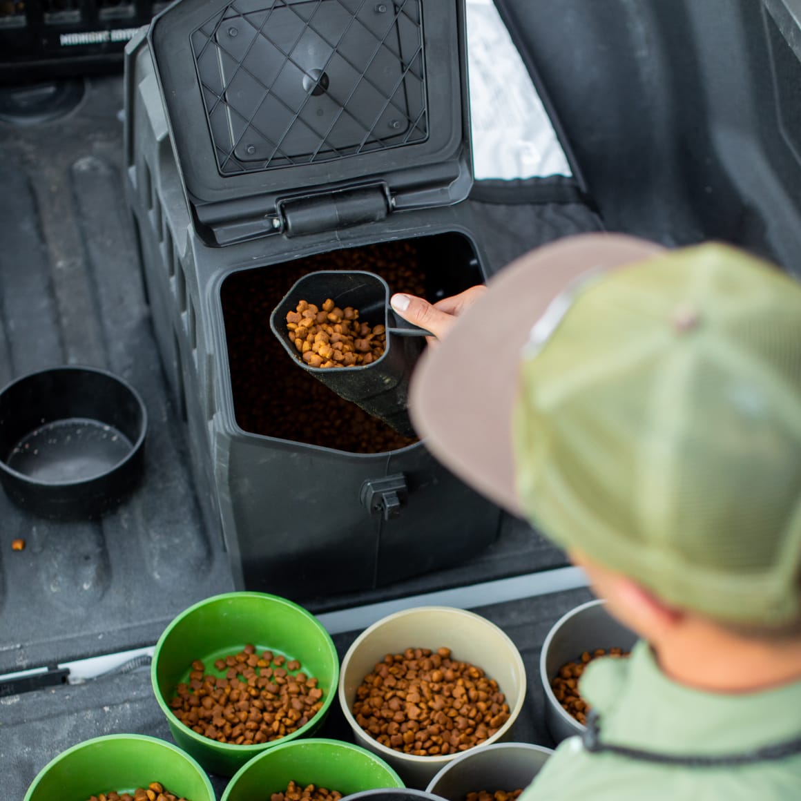 A person uses a scoop to fill several green and gray bowls with dry pet food from a GUNNER FOOD CRATE - Black by GUNNER, viewed from above.