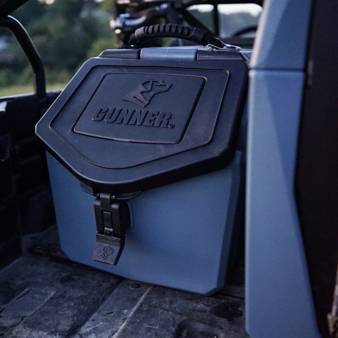 A GUNNER FOOD CRATE - Ducks Unlimited Blue rests on a vehicle’s backseat, partly illuminated, with a blurred forest backdrop—showcasing the quality and craftsmanship you expect from the American-made GUNNER brand.