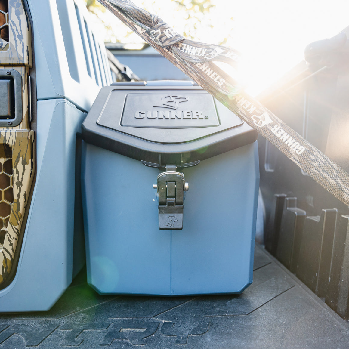A GUNNER FOOD CRATE - Ducks Unlimited Blue with a black lid is secured in a vehicle next to a patterned container. Sunlight highlights the latch and the GUNNER-branded strap above.