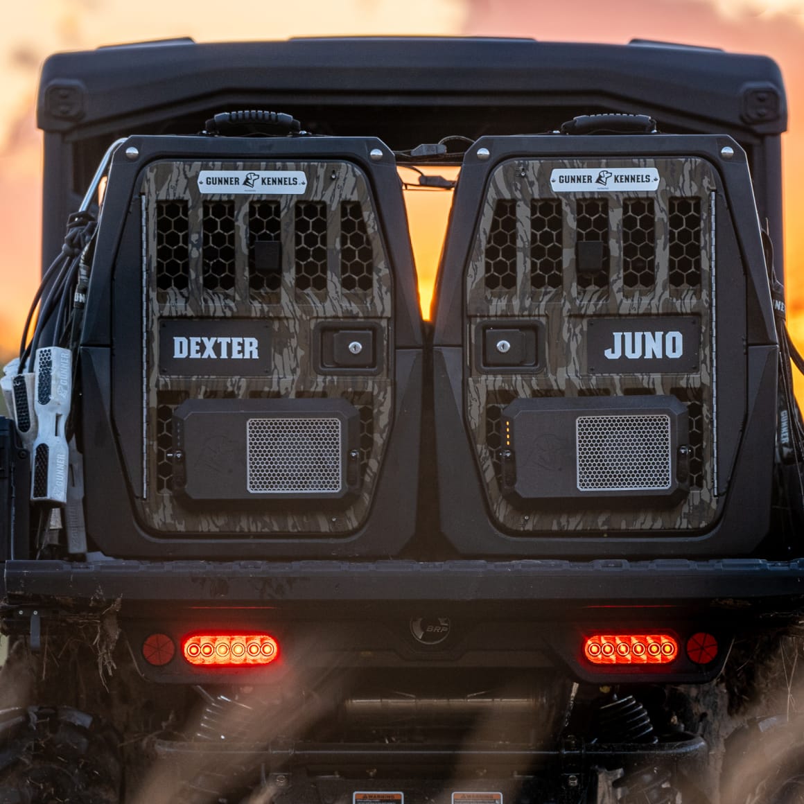 Two GUNNER G1™ KENNEL - MOSSY OAK BOTTOMLAND BLACK crates, featuring a camo pattern and mesh doors, are secured in the back of a vehicle at sunset as red brake lights glow beneath them.