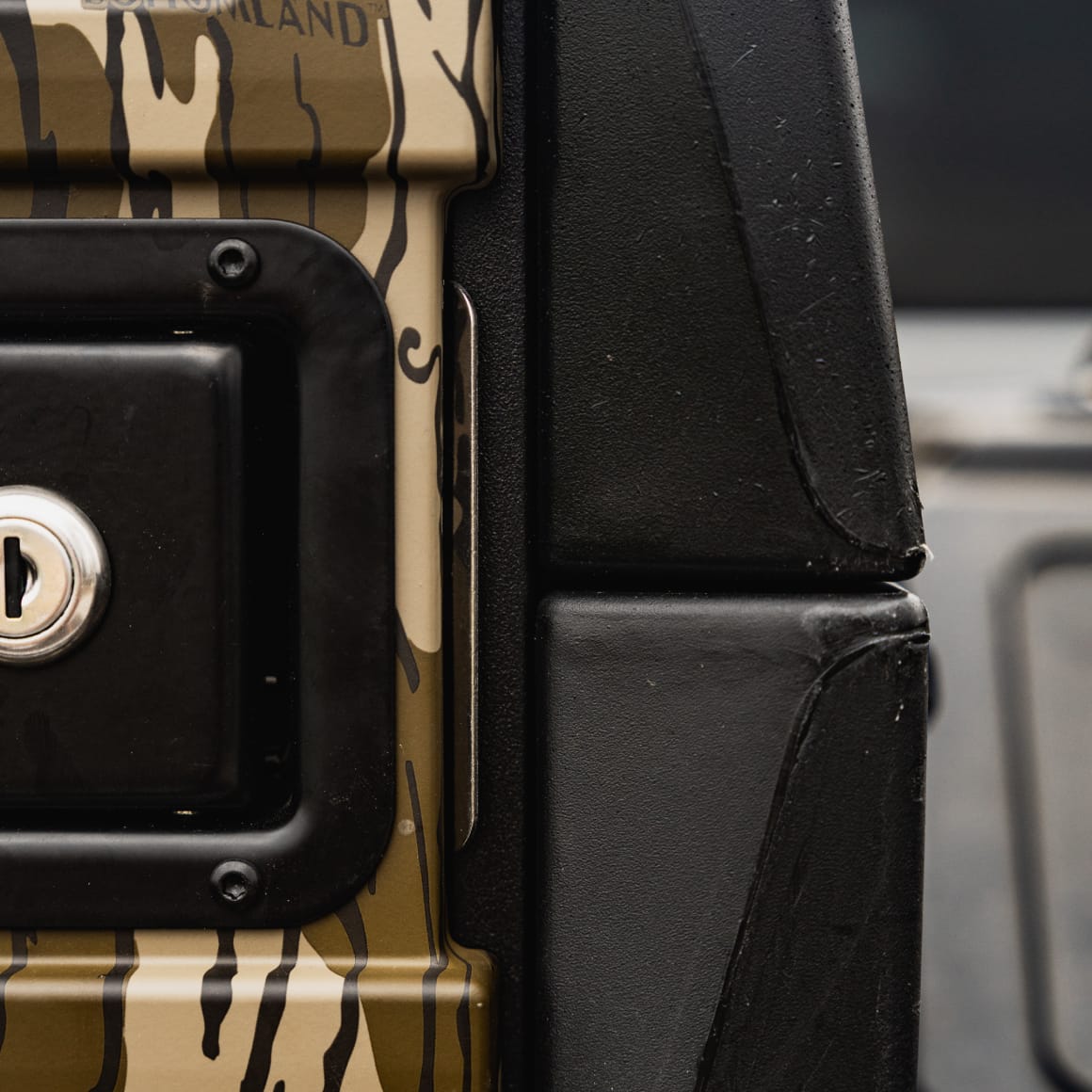 Close-up of a vehicle door with camouflage, black lock, and keyhole beside textured black surface—perfect for securely installing the GUNNER G1™ KENNEL - MOSSY OAK BOTTOMLAND BLACK to keep your pet safe during travel.