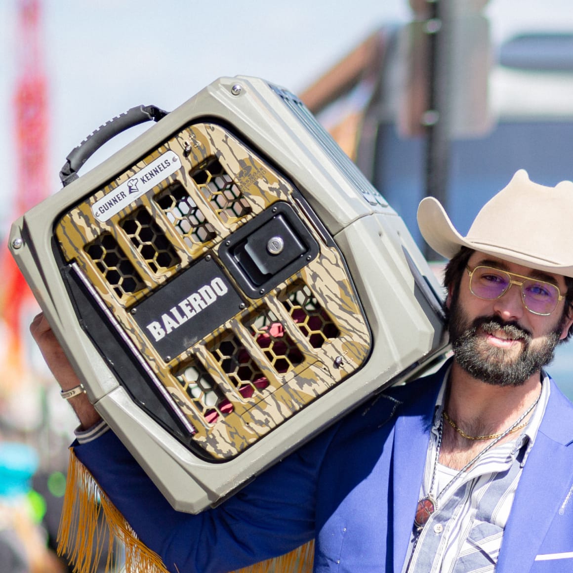 A man in a tan cowboy hat, sunglasses, and blue suit jacket carries a durable GUNNER G1™ KENNEL - MOSSY OAK BOTTOMLAND GUNMETAL on his shoulder at an outdoor event.
