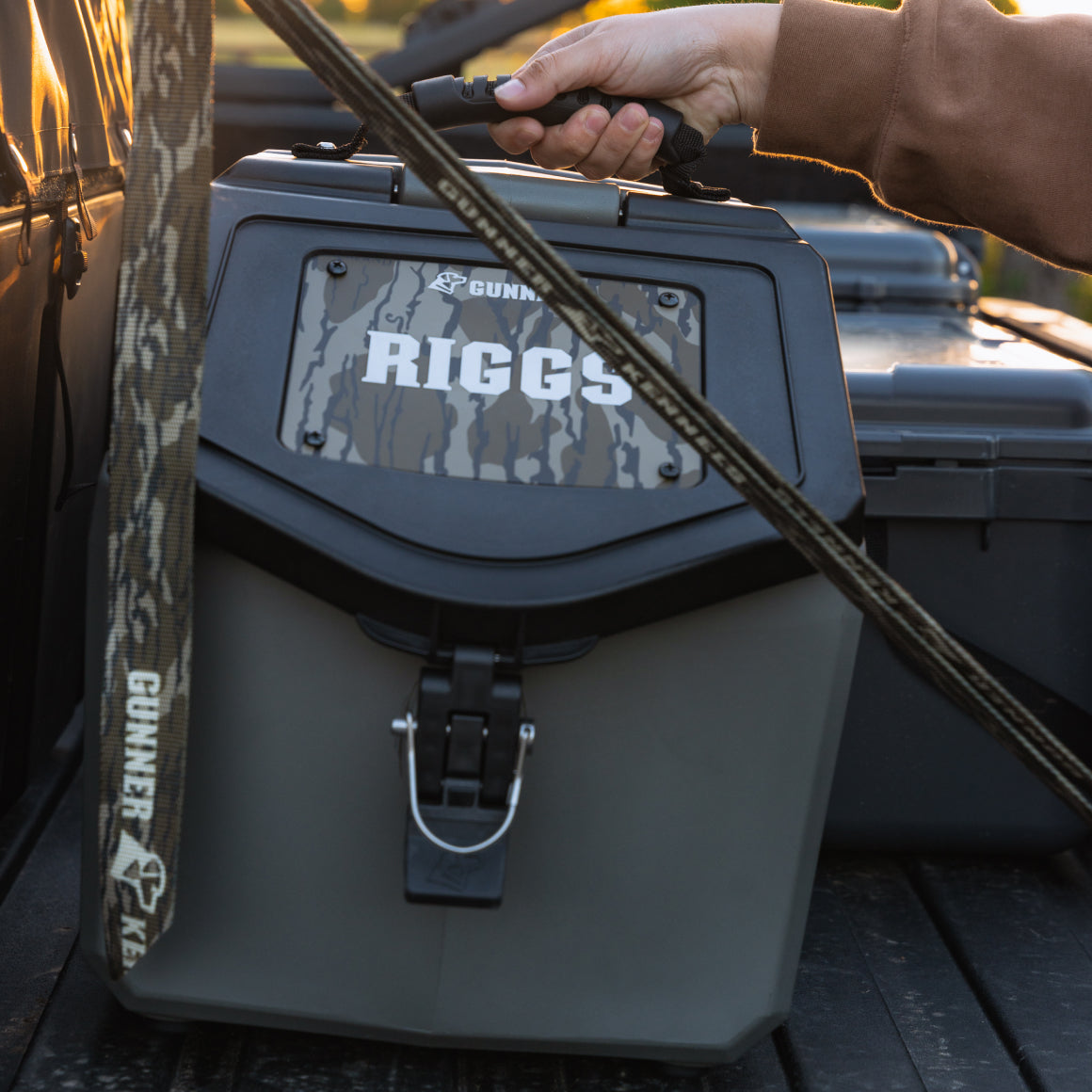 A hand lifts the GUNNER FOOD CRATE - GUNMETAL, a sturdy, camo-patterned waterproof dog food container with the GUNNER logo, secured in a truck bed. A camouflage strap with GUNNER text is visible in the foreground.