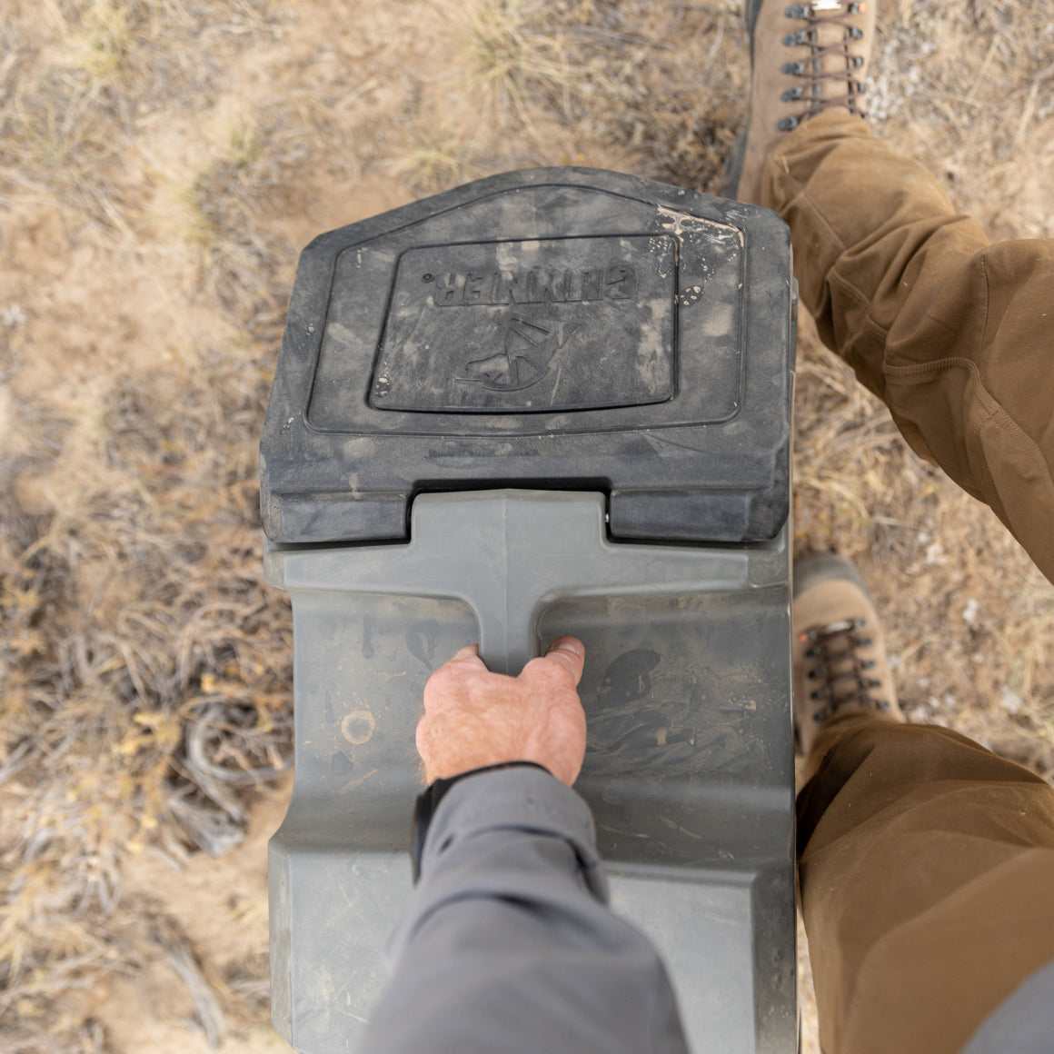 A person in brown boots and pants holds the handle of a GUNNER FOOD CRATE - GUNMETAL by GUNNER while standing on dry, grassy ground outdoors.