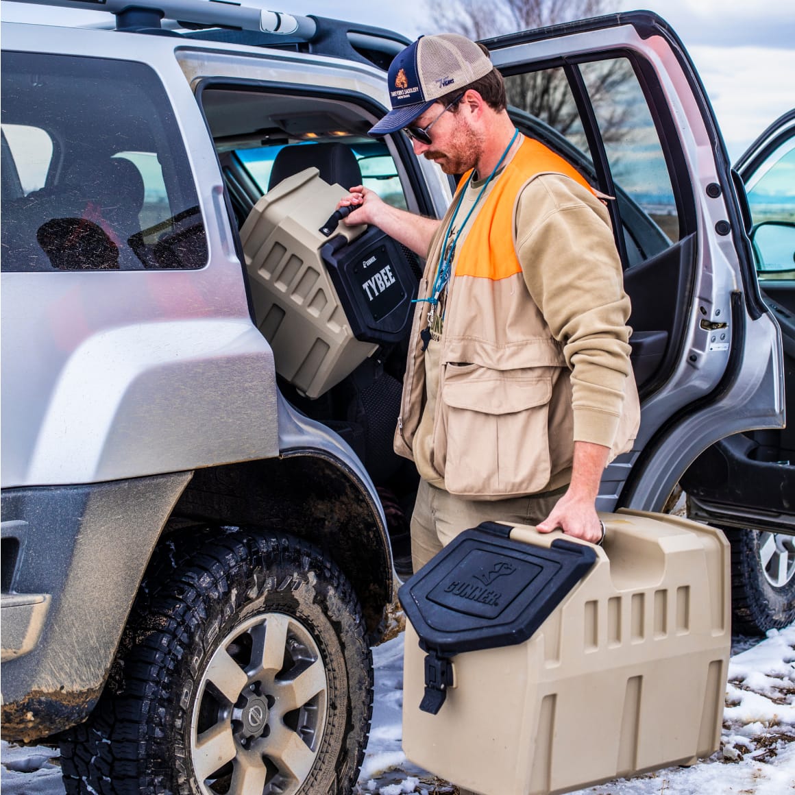 A man in outdoor gear loads a GUNNER FOOD CRATE - TAN by GUNNER and beige and black storage bins into the back seat of a muddy SUV parked on snowy ground.