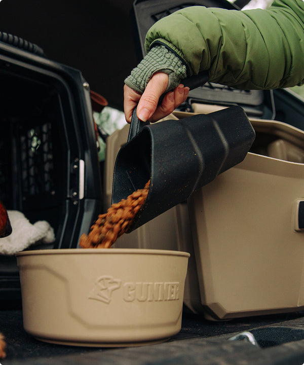 A person in a green jacket pours dry dog food from a black scoop into a beige bowl labeled GUNNER, with a dog kennel and pet supplies visible near the crate in the background.
