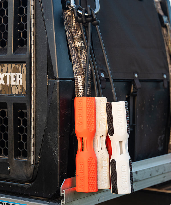 Three rubber dog training dummies—orange, white, and black and white—hang by straps from a rugged vehicle equipped with a dog crate, black cage, and camouflage detailing—essential outdoor gear for any hunting dog.