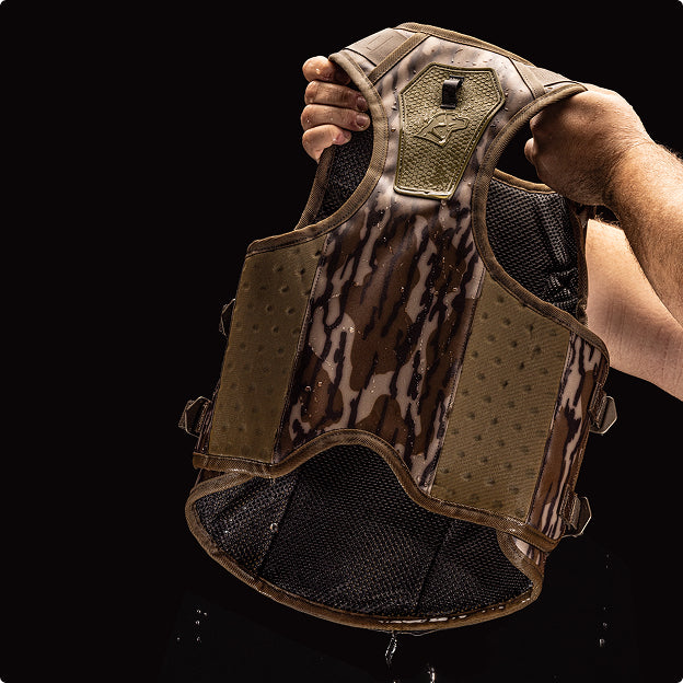 A person holds up a camouflage-patterned tactical vest with mesh lining, ideal for outfitting a working dog, displayed against a black background.