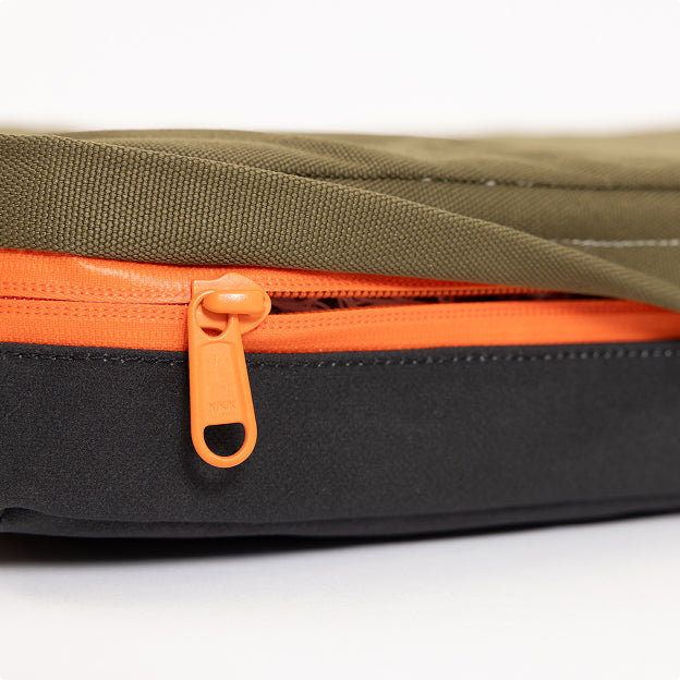 Close-up of an orange zipper on a bag with olive green fabric on top and black fabric on the bottom, designed for outdoor gear, against a white background.