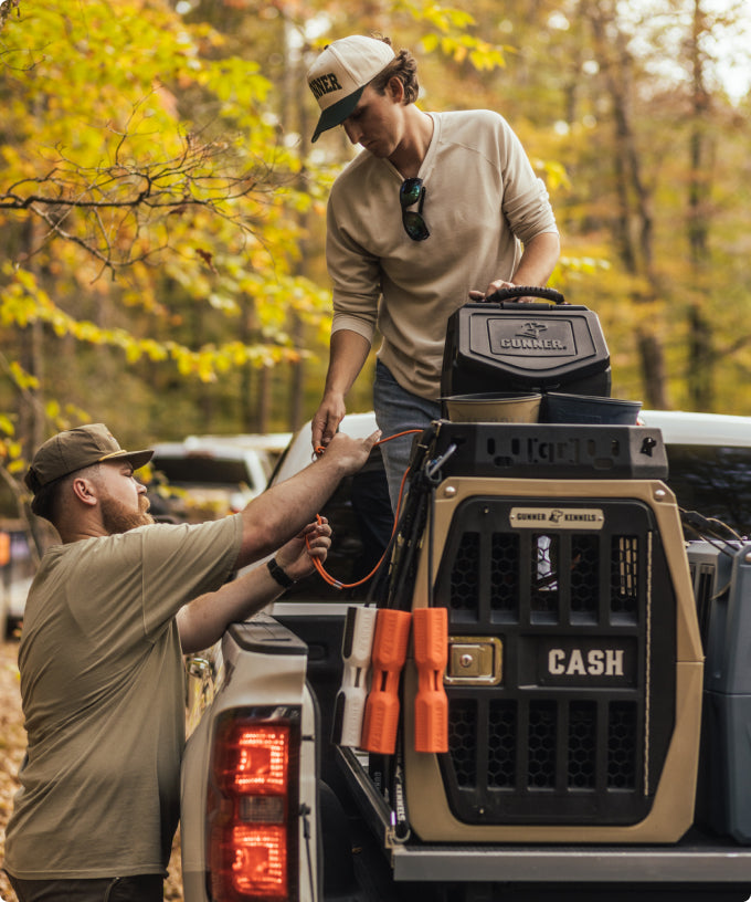 Two men stand at the back of a pickup truck in a wooded area, handing an item to each other. The truck bed holds a kennel labeled CASH with orange training dummies attached, likely for a hunting dog. Autumn leaves add color to the outdoor scene.
