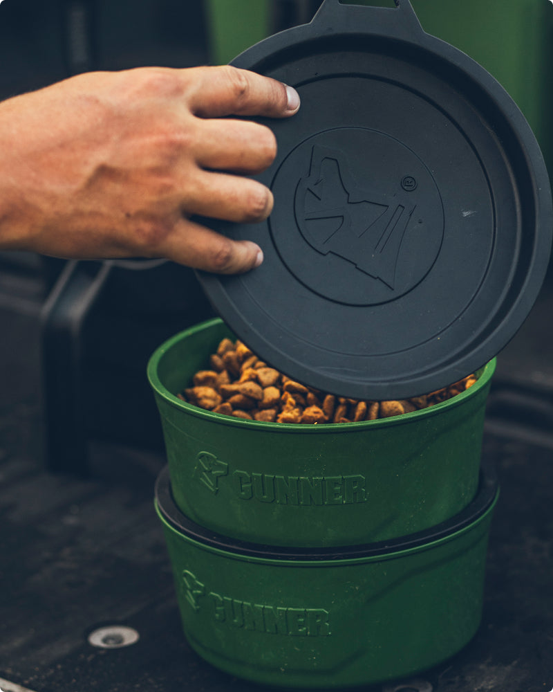 A hand lifts the lid off a stackable green dog food container labeled GUNNER, featuring a dog logo—perfect for storing kibble for your hunting dog or working dog.