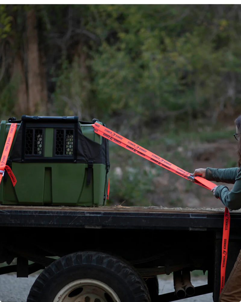 A person secures a green dog kennel with orange tie-down straps on the bed of a flatbed truck, surrounded by a natural outdoor setting with trees in the background.