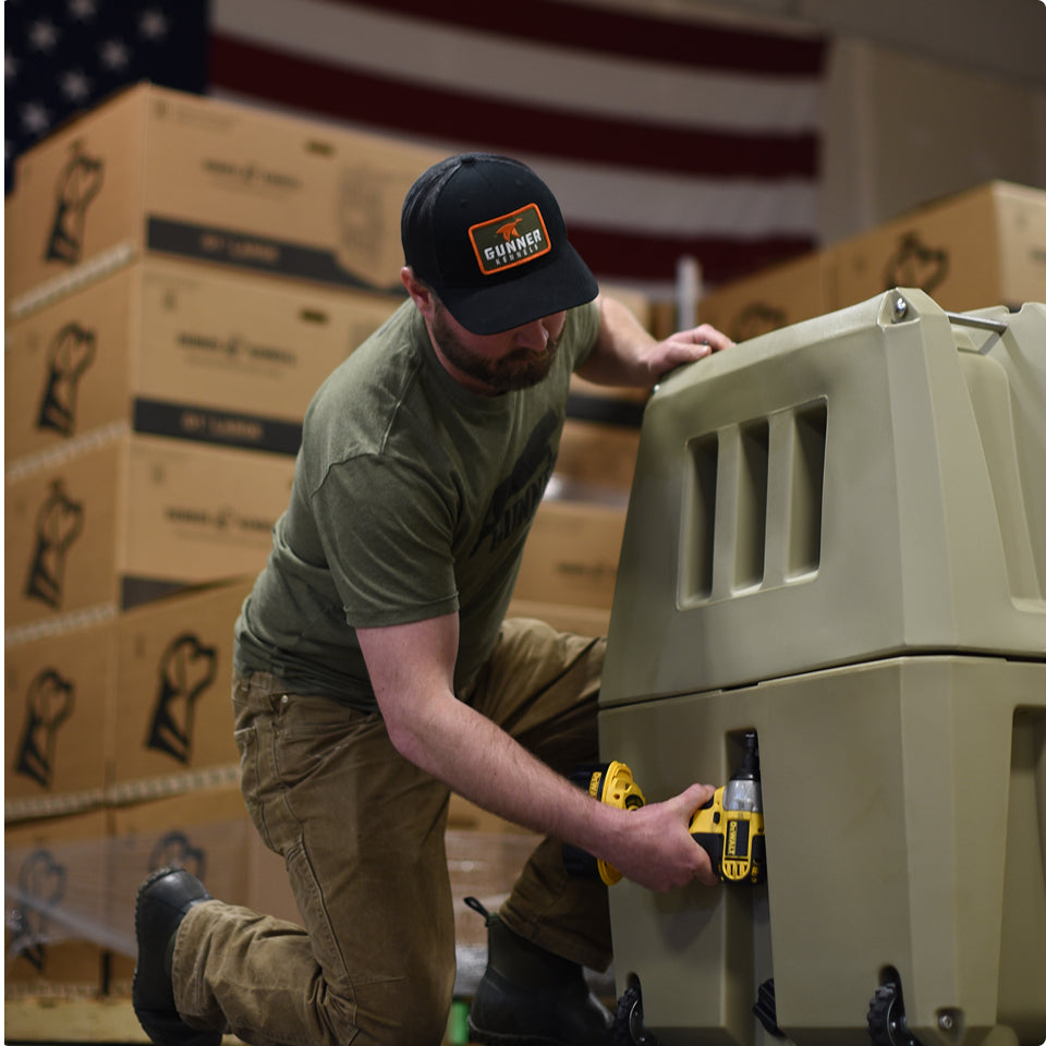 A man wearing a cap and green t-shirt kneels while using a yellow power drill to assemble a large tan dog kennel. Stacked boxes and a U.S. flag are visible in the background.