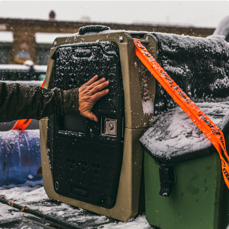 A person’s hand rests on a large, snow-covered dog kennel secured with orange straps in the back of a vehicle. Snow blankets the kennel, nearby bins, and surrounding area.