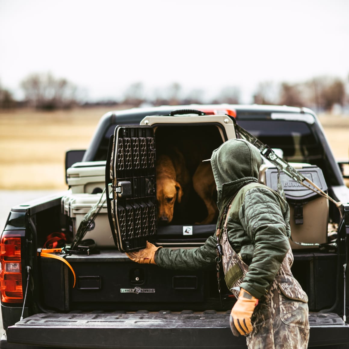 A person in outdoor gear opens the GUNNER G1™ KENNEL - MOSSY OAK BOTTOMLAND TAN in the back of a pickup, releasing a brown dog. The truck is parked in a rural area with fields and trees in the background.