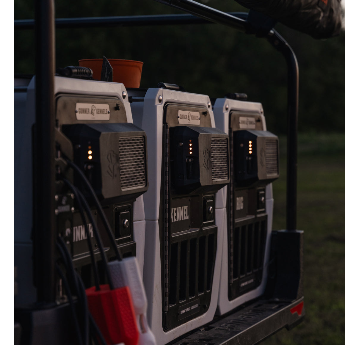 Three portable GUNNER Kennels are secured on a vehicle platform outdoors, each equipped with ventilation panels and the GUNNER FAN KIT. Cables and clamps connect the kennels to power sources, highlighting essential GUNNER accessories.