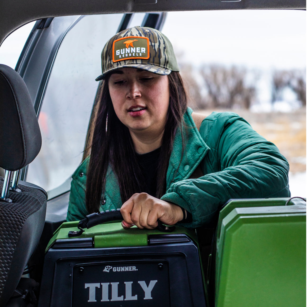 A woman in a green jacket and GUNNER BOTTOMLAND MALLARD TRUCKER hat loads a black TILLY pet carrier into her vehicle, with a snowy landscape visible through the open door.