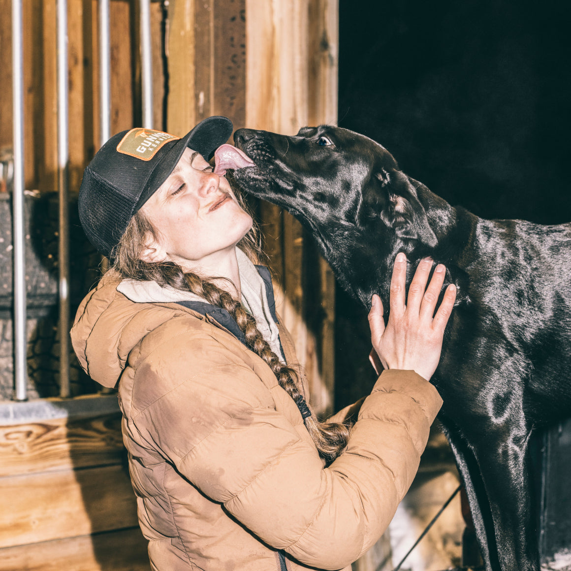 Wearing a GUNNER BLACK MALLARD TRUCKER hat and a brown jacket, a woman smiles as a large black dog licks her face. She gently holds the dogs neck outside near a wooden structure at night.