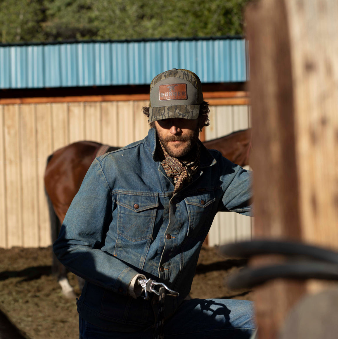 A man wearing the GUNNER BOTTOMLAND HAT and a denim jacket stands by a wooden fence, with a brown horse and metal barn roof in the background, appearing to handle ropes or reins.