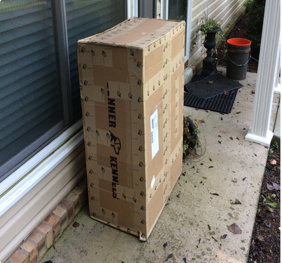 A large cardboard box labeled Inner Kennels sits on a concrete porch next to a house, near a window and some garden items, including an orange bucket and outdoor gear for a working dog.