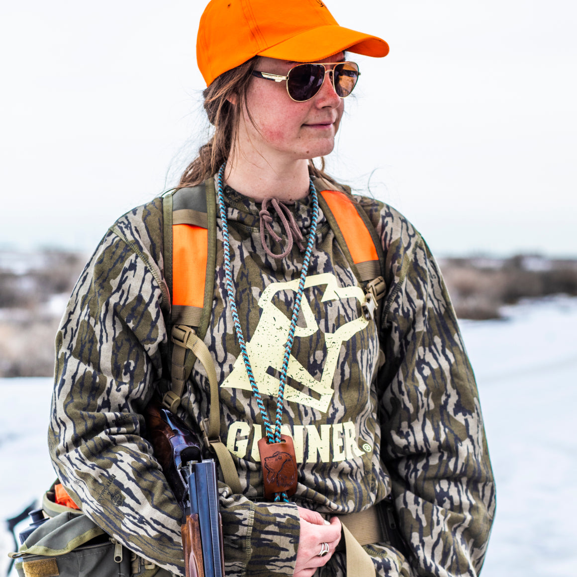 A woman stands outdoors with a shotgun resting in her arm, wearing the GUNNER BOTTOMLAND HOODIE, bright orange cap, and sunglasses. Snow and dry brush can be seen in the background.