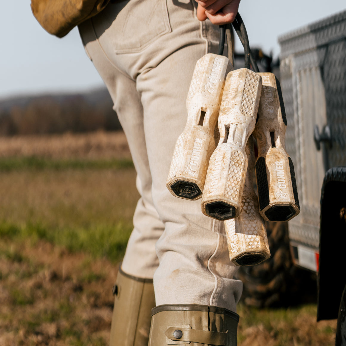 Wearing beige pants and green boots, a person stands near a metal trailer in a grassy rural area, holding several GUNNER BUMPER training dummies from GUNNER for effective outdoor training.