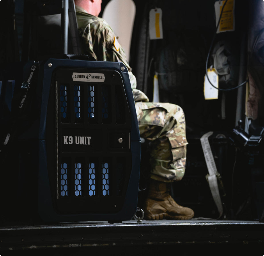 A person in military uniform sits in a vehicle next to a secured black dog crate labeled K9 UNIT with blue vents and a GUNNER KENNELS sign on top, showcasing essential outdoor gear for any hunting dog.