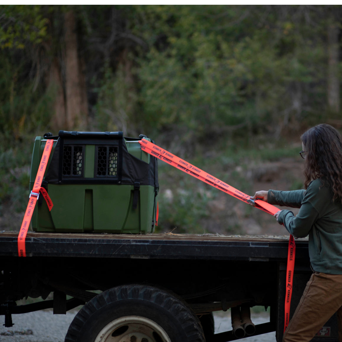 A person secures a green, crash test certified dog crate on the back of a flatbed truck outdoors using bright orange GUNNER TIE-DOWN STRAP KIT, surrounded by trees and greenery.