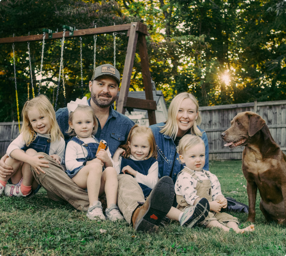 A family of five with three young children and a brown dog sit on grass in a backyard near a swing set, smiling at the camera; a dog kennel is visible by the trees and wooden fence in the background.